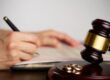 Close-up of a person signing divorce document, with a judge gavel and wedding rings in focus.
