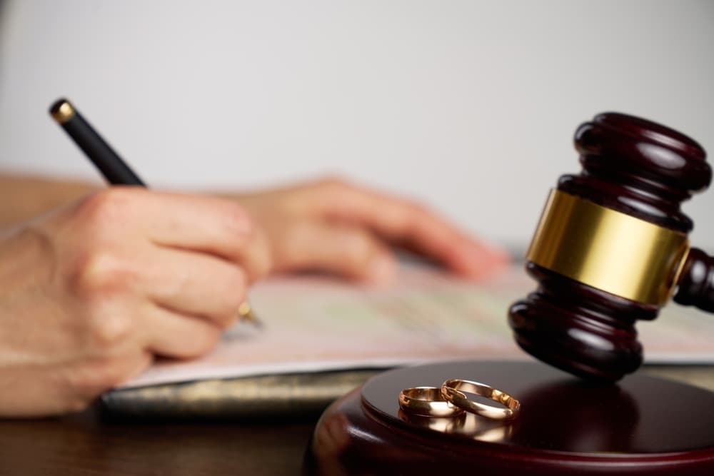 Close-up of a person signing divorce document, with a judge gavel and wedding rings in focus.